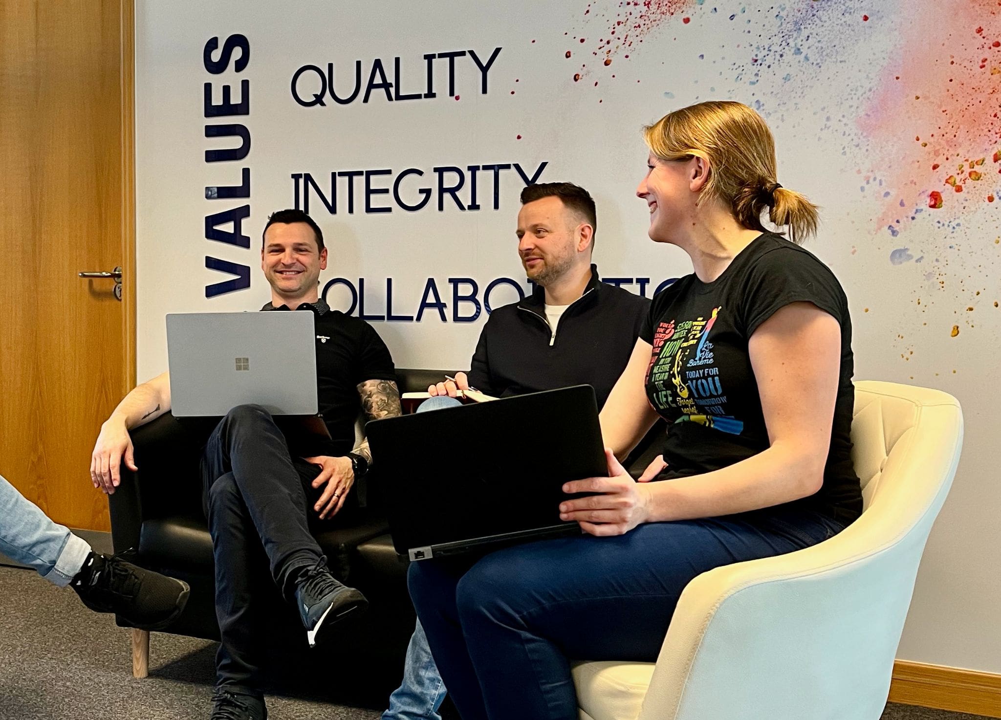 Bob Simon and louise all sat on a chair with their laptops infront of the mexa solutions logo
